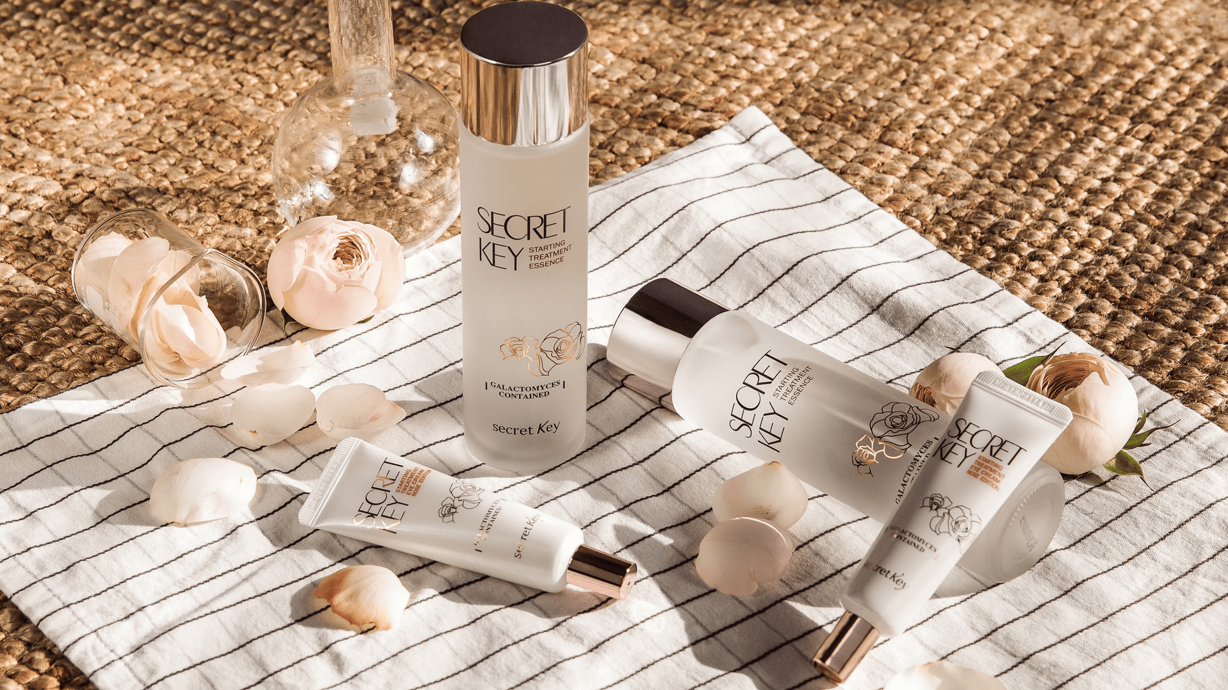 Secret Key Starting Treatment Essence och hudvårdsprodukter placerade på randig duk med rosenblad och glasdetaljer i en estetisk produktuppställning.