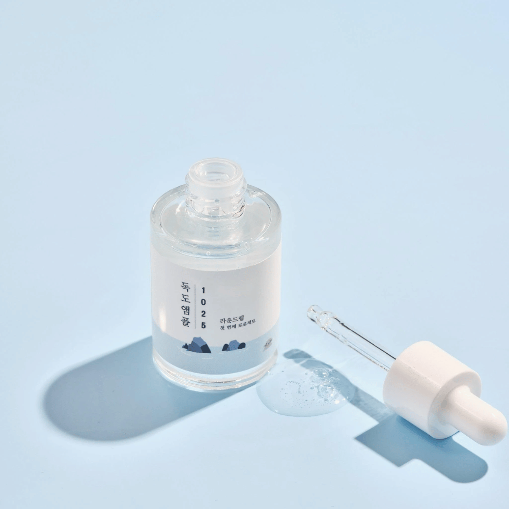 An open bottle of 1025 Dokdo Ampoule with a dropper next to it, against a blue background creating a soft shadow.