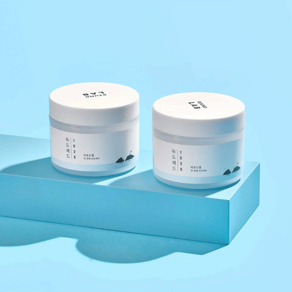 Two white jars of 1025 Dokdo Pads, placed on a light blue shelf casting a reflection, against a blue background.