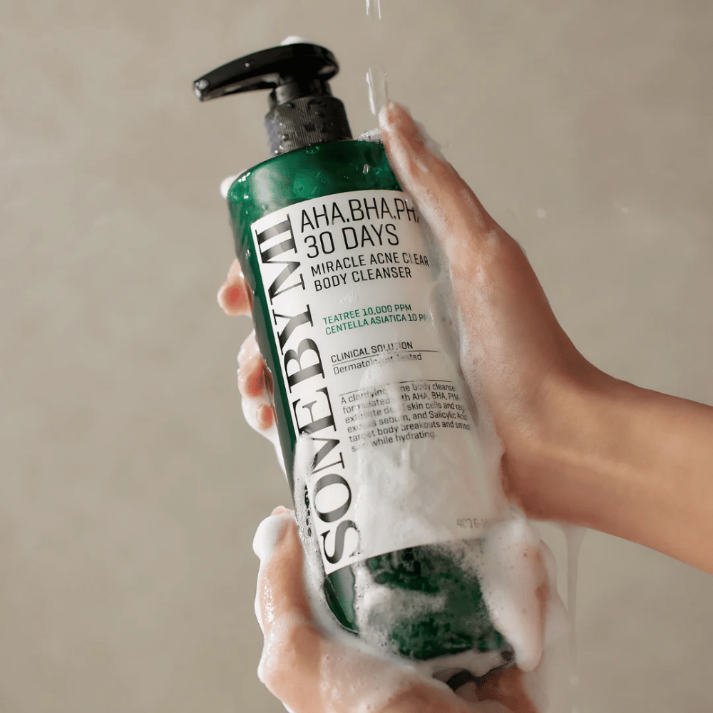 A person is holding a bottle of SOME BY MI AHA.BHA.PHA 30 Days Miracle Acne Clear Body Cleanser. The bottle is green with a white label and is covered in foam from the cleanser. The person is washing with the product while water runs over their hands.