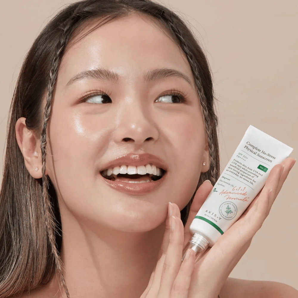 The picture shows a young woman smiling happily and holding a tube of AXIS-Y Complete No-Stress Physical Sunscreen. She has a radiant and natural appearance with minimal makeup, which highlights the product she is presenting.