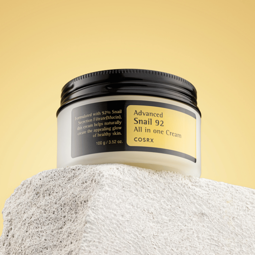 A jar of COSRX Advanced Snail 92 All in one Cream placed on a rocky surface with a yellow background.
