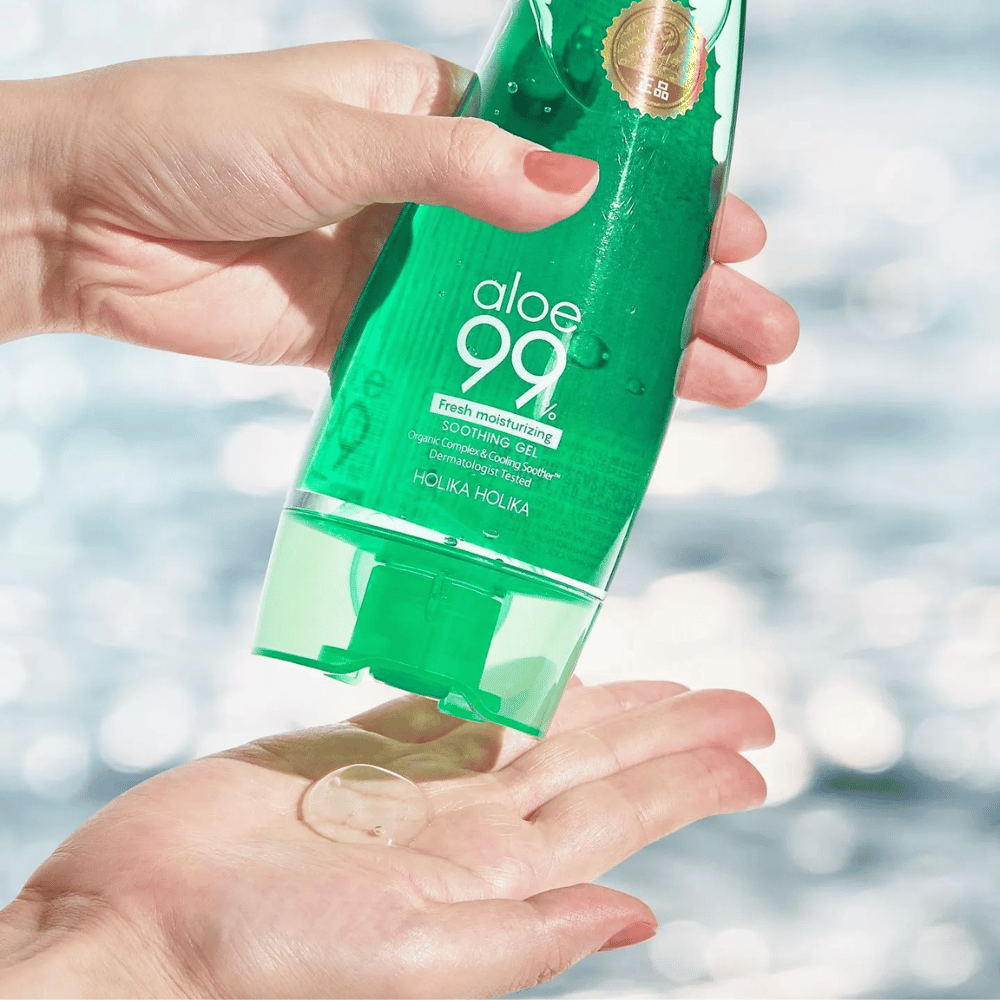 A hand holds a green tube with Holika Holika Aloe 99% Soothing Gel, while another hand catches a dollop of gel being dispensed. The background is blurry and shows a watery, sparkling pattern.