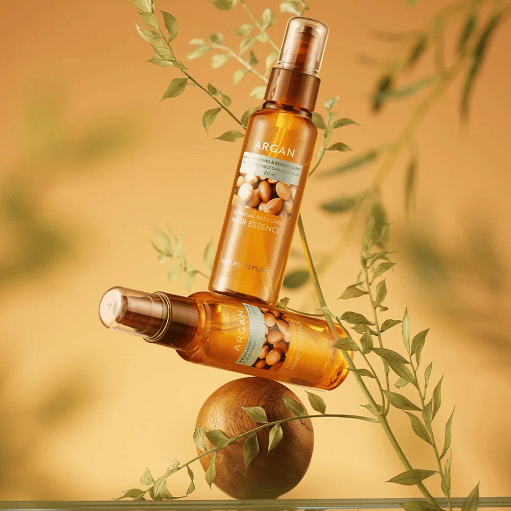 Two bottles Nature Republic Argan Essential Moisture Hair Oil placed among green leaves – silky shine for dry hair.