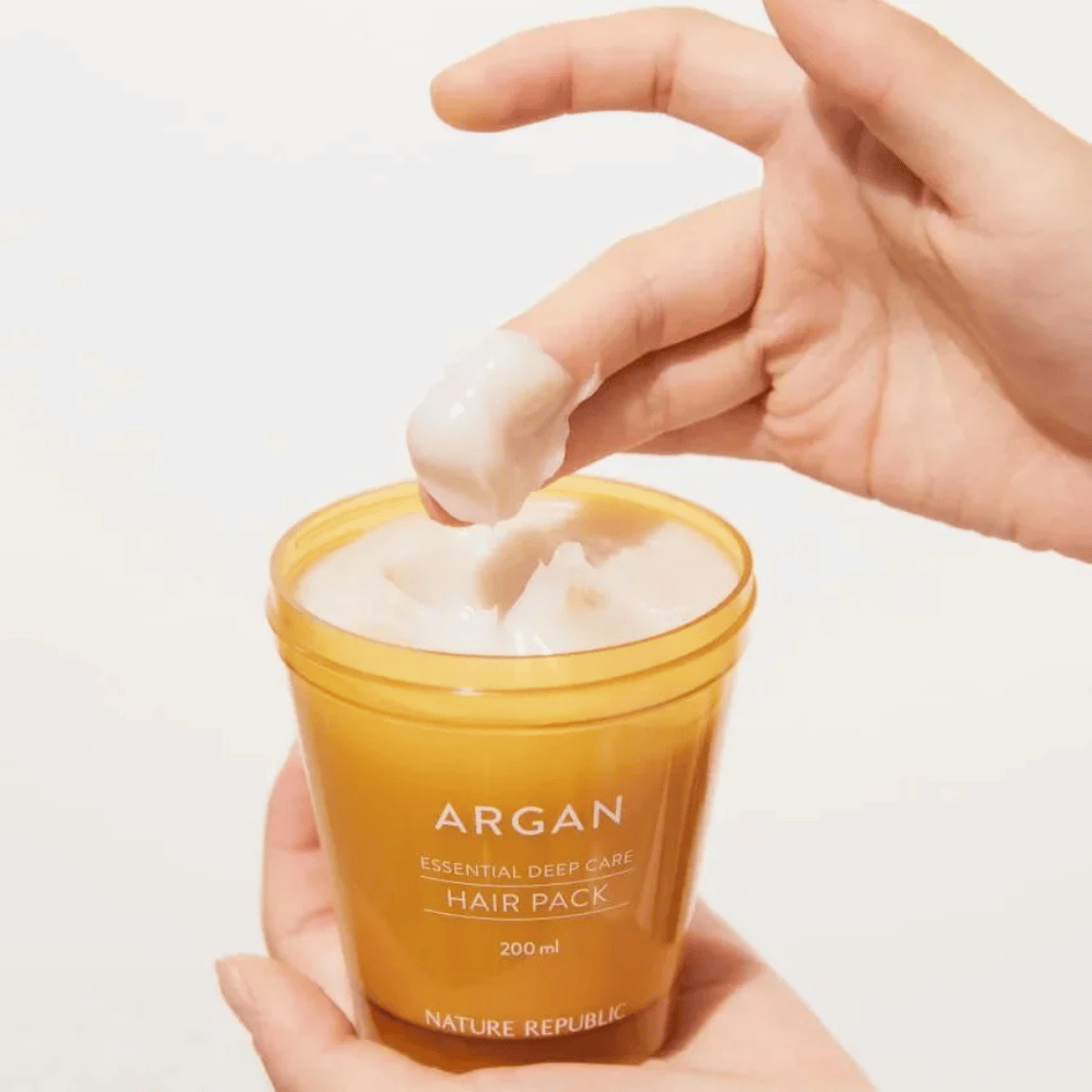 Hand picks up the creamy formula from Nature Republic Argan Essential Moisture Repairing Hair Mask – intensive nourishment for dry hair.