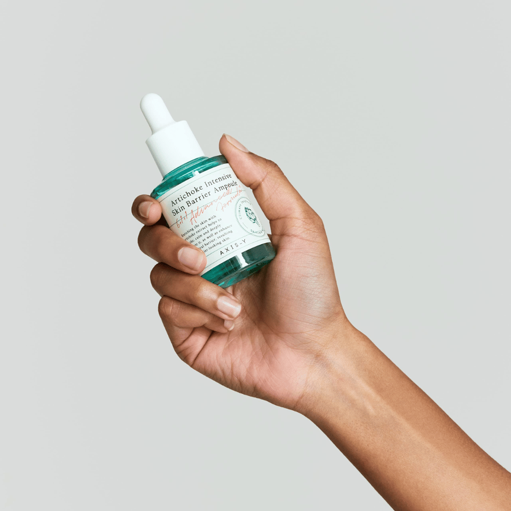 A hand holds a bottle of skincare serum with the label "Artichoke Intensive Skin Barrier Ampoule.