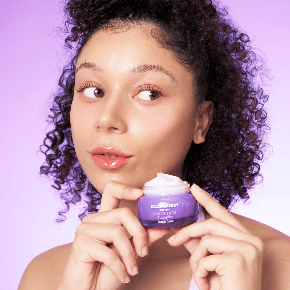 Woman holds EQQUALBERRY Bakuchiol Plumping Capsule Cream in her hand and shows off her hydrated, glowing skin against a purple background.