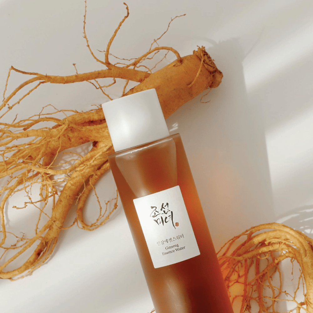 A brown bottle of Beauty of Joseon Ginseng Essence Water with a white cap lies next to a fresh ginseng root on a white background.