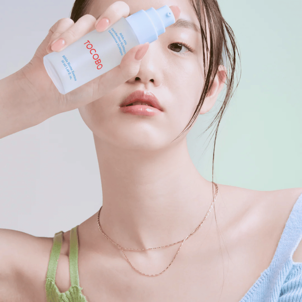 Young woman with soft makeup and natural skin tone holds a TOCOBO Bifida Biome Essence bottle in front of her face. She is wearing a light blue sweater with green shoulder straps and a thin gold necklace against a light background.