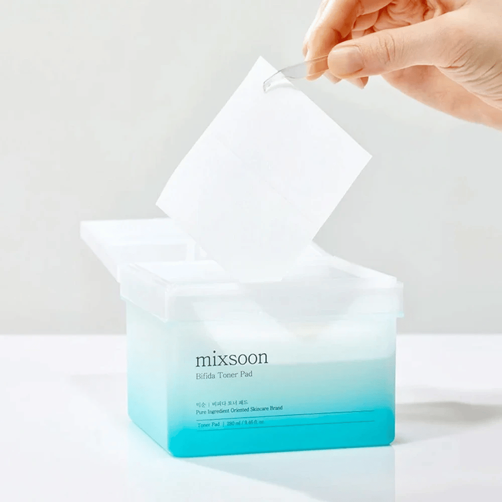 A transparent container with a blue and white gradient containing Bifida toner pads. A hand takes a toner pad from the container, which is labeled with "mixsoon Bifida Toner Pad" and holds 280 ml (9.46 fl. oz.) of toner pads intended for skincare.