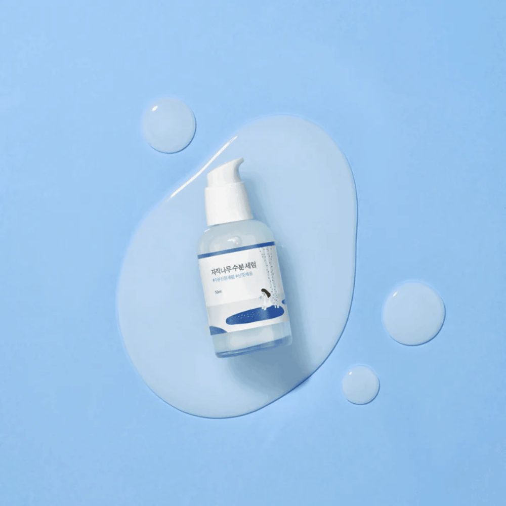 Round Lab Birch Juice Moisturizing Serum in a transparent pump bottle placed on a blue background with drops and spilled serum.