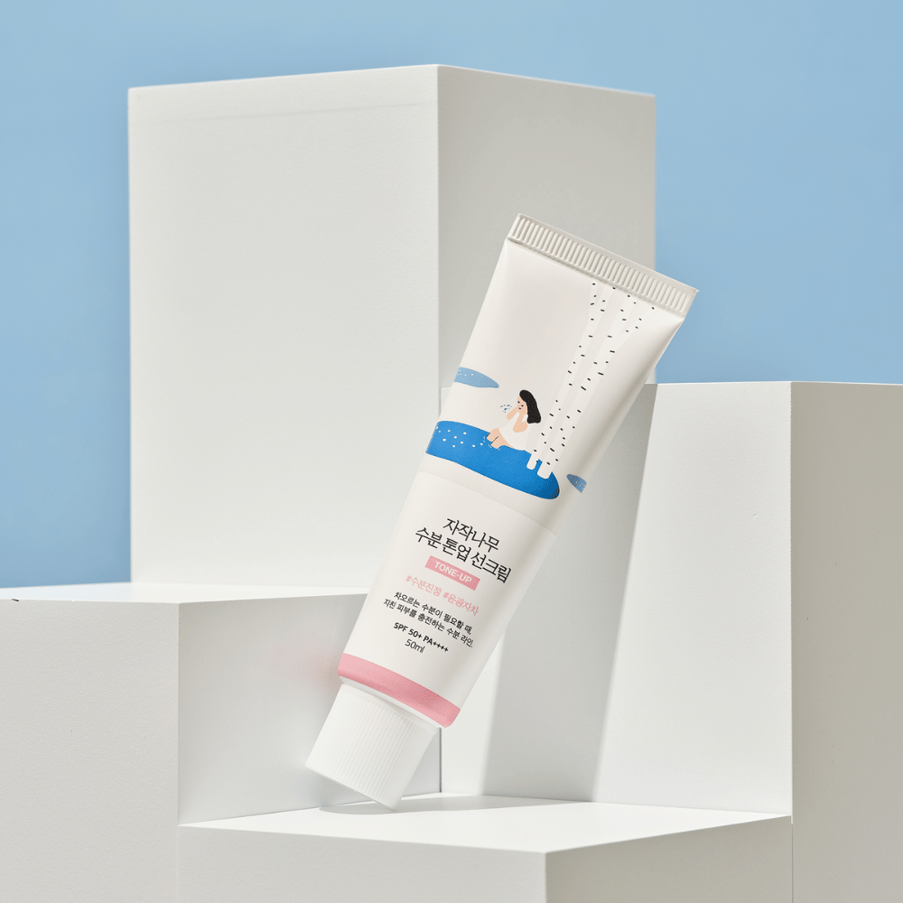 A tube with Birch Juice Moisturizing Tone Up Sunscreen placed on white cubes against a light blue background. The tube is white with an illustration of a woman by a birch tree and water. The text in Korean indicates that the product has SPF 50+ PA++++ and contains 50 ml.