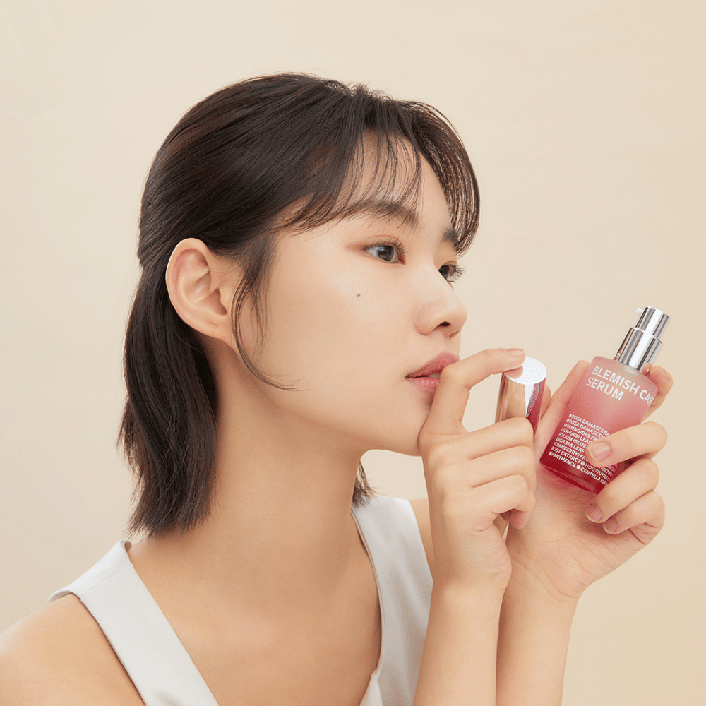 A woman with dark hair holds a bottle of Blemish Care Serum in one hand and removes the cap with the other. She has a neutral facial expression and the background is light beige, giving a soft and minimalist impression.