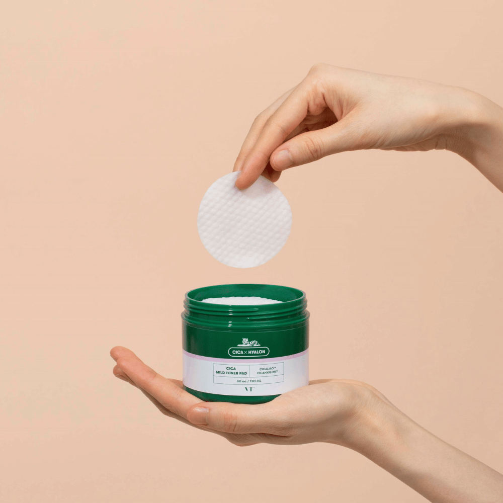 Hand holding a green jar with VT Cica Mild Toner Pad and lifting a round, white toner pad over the jar against a beige background.