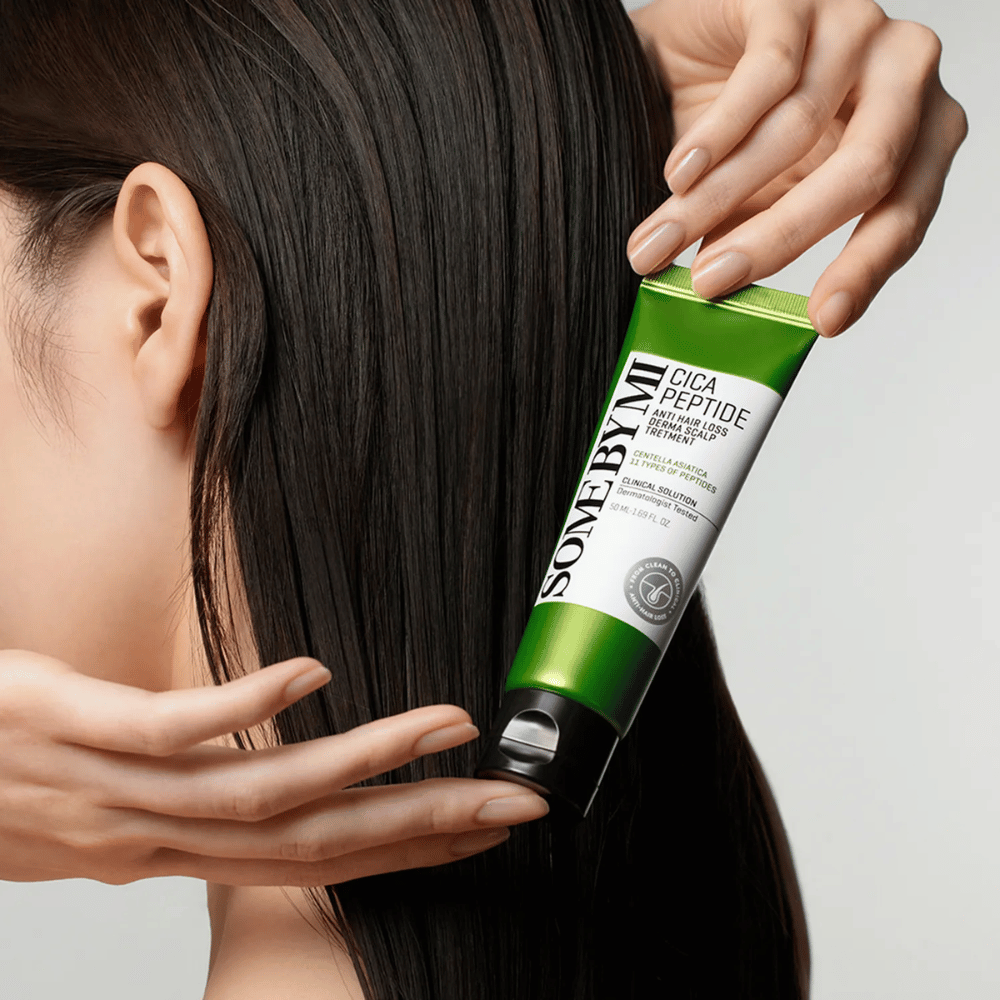 Woman applies SOME BY MI Cica Peptide Anti Hair Loss Derma Scalp Treatment in long, dark hair with a green and white tube.