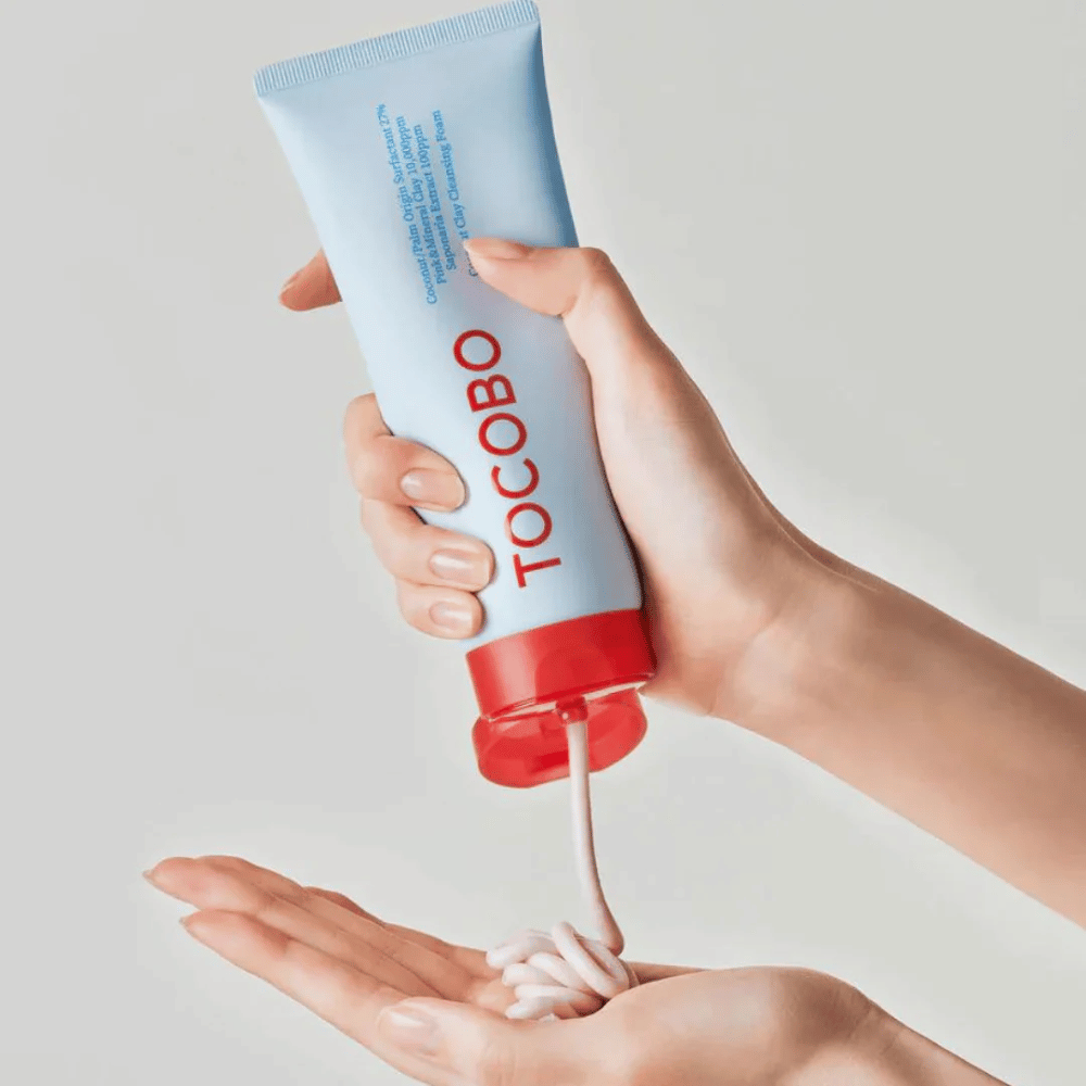 A person is holding a tube of TOCOBO Coconut Clay Cleansing Foam. The tube is light blue with a red cap and the text "TOCOBO" on the front. The product is squeezed out of the tube and shaped like a shampoo in the hand. The background is neutral, drawing focus to the product.