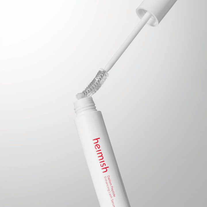 Close-up of the applicator for Heimish Dailism Peptide Enhancing Lash Serum. The brush is pulled up from the white container and shows the transparent serum formula that is applied to the lashes.