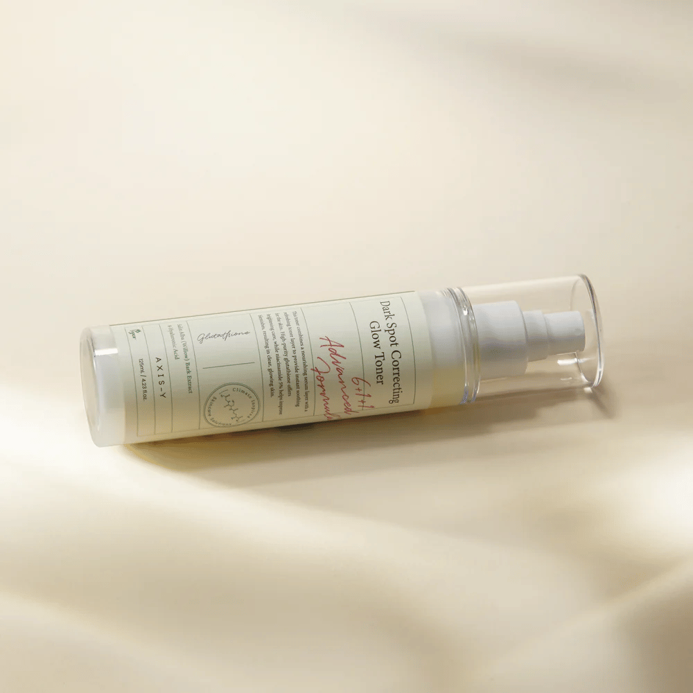 A lying bottle of "AXIS-Y Dark Spot Correcting Glow Toner" placed on a light surface. The bottle is transparent with a white label and has a white spray pump. The label shows information about the product's brightening and moisturizing properties and contains key ingredients such as Glutathione and Niacinamide.