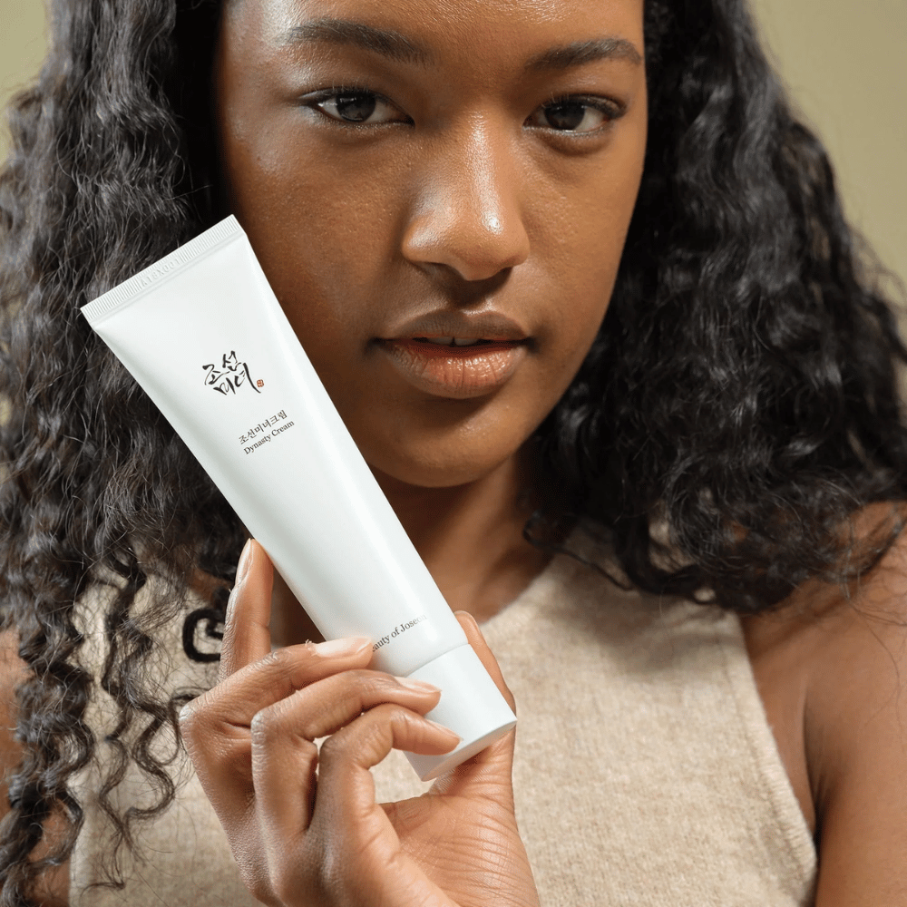 A woman with curly hair holds a tube "Beauty of Joseon Dynasty Cream" close to her face, highlighting the product. The cream, presented in a sleek white tube, is designed to nourish and rejuvenate the skin, emphasizing its appeal for different skin types.