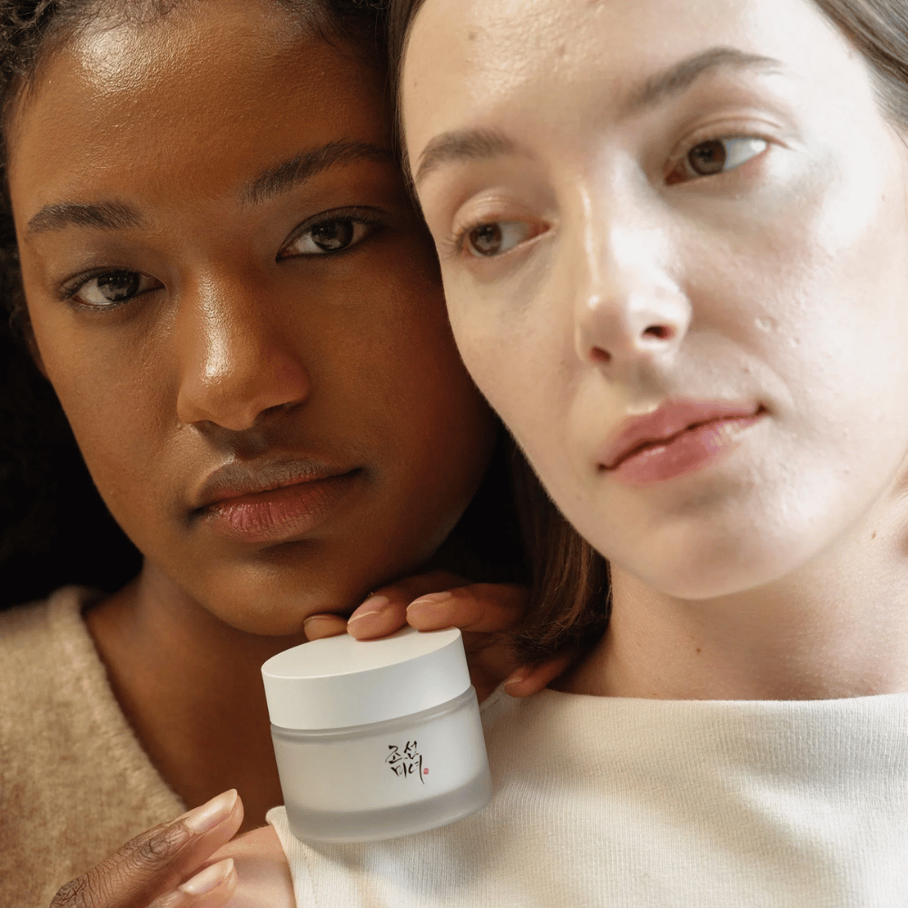 Two women, one with a darker and one with a lighter skin tone, stand close together focusing on their faces and a skincare cream from "Beauty of Joseon". The cream is placed in front of them, emphasizing its use for different skin types and its ability to provide glow and nourishment.