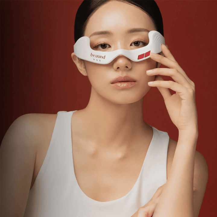 Model image where a person is using Beaund Eye Rejuv, a futuristic LED device for the area around the eyes, against an elegant red background.