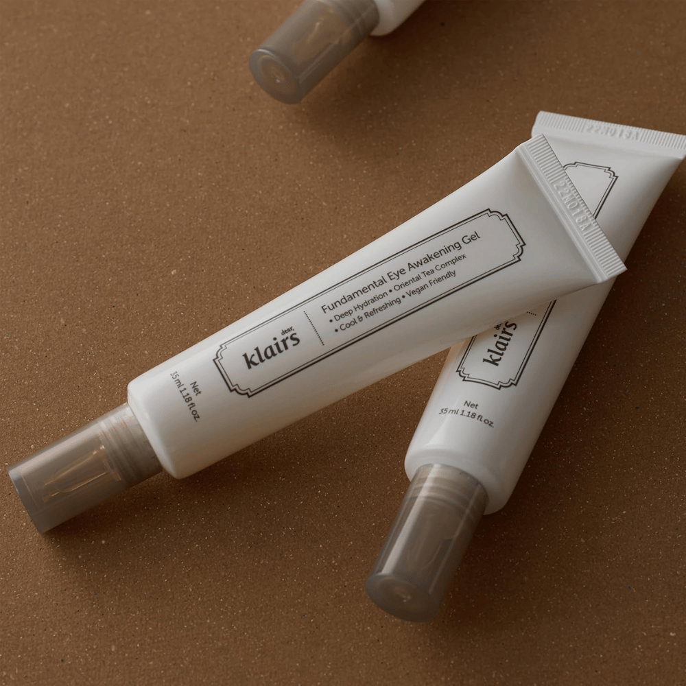 Lifestyle image with two tubes of Klairs Fundamental Eye Awakening Gel placed against a warm brown background. The composition highlights the product’s elegant design and premium feel while keeping the focus on eye care and hydration.