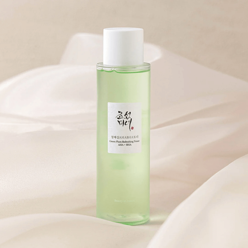 A bottle of "Green Plum Refreshing Toner" AHA + BHA stands on a soft, light background. The product has a green liquid that reflects freshness and naturalness. The label is white with black and red accents, giving an elegant and clean look.