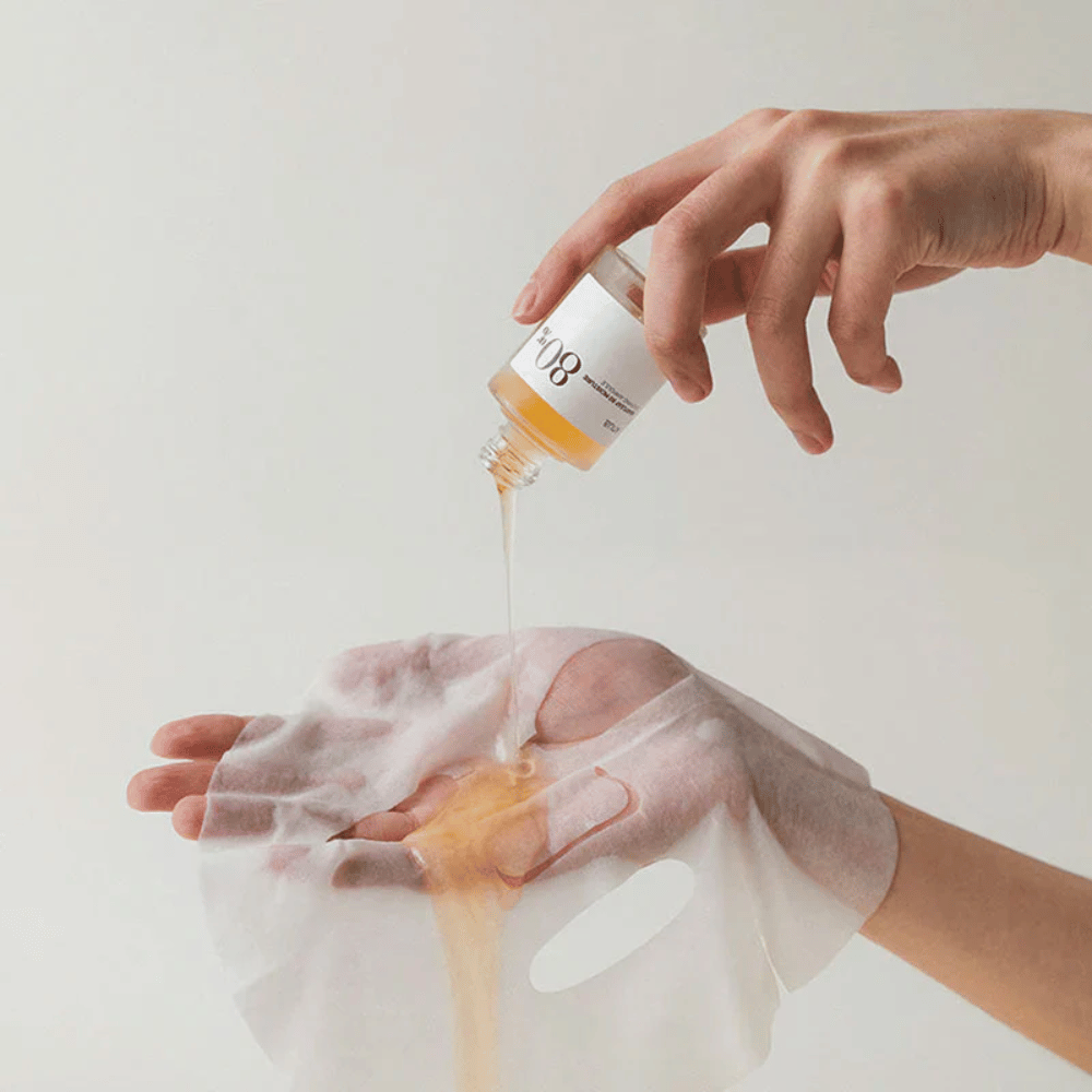 A hand pours a golden, thick liquid from a small bottle labeled '108' onto a transparent face mask held in another hand. The background is neutral and uniform to highlight the product.