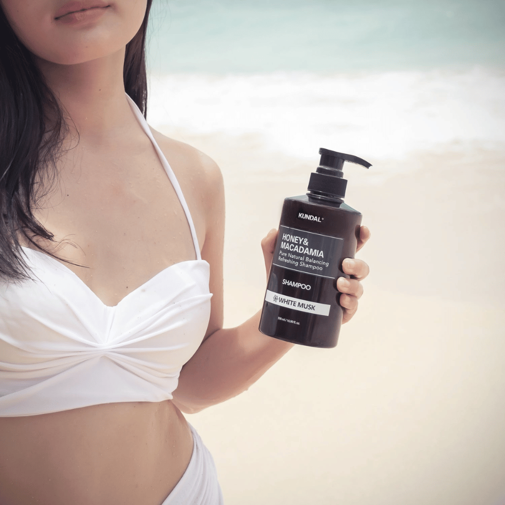Woman in white bikini holds KUNDAL Honey & Macadamia Nature Shampoo White Musk on a beach with the sea in the background.