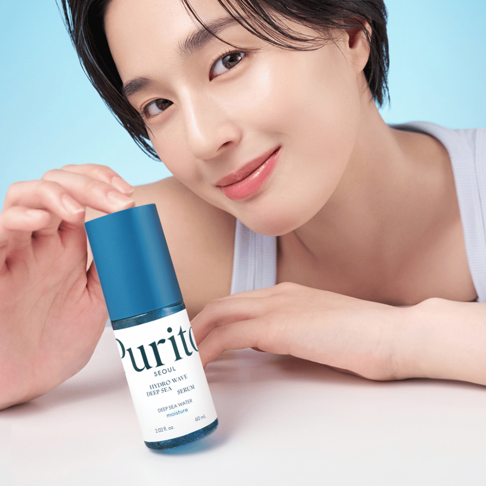 A woman with short hair holds a blue serum from Purito, Hydro Wave Deep Sea Serum. Her skin looks radiant and moist, highlighting the product in focus. The background is light blue, giving a fresh and clean feeling. The product is immersed in skin care.