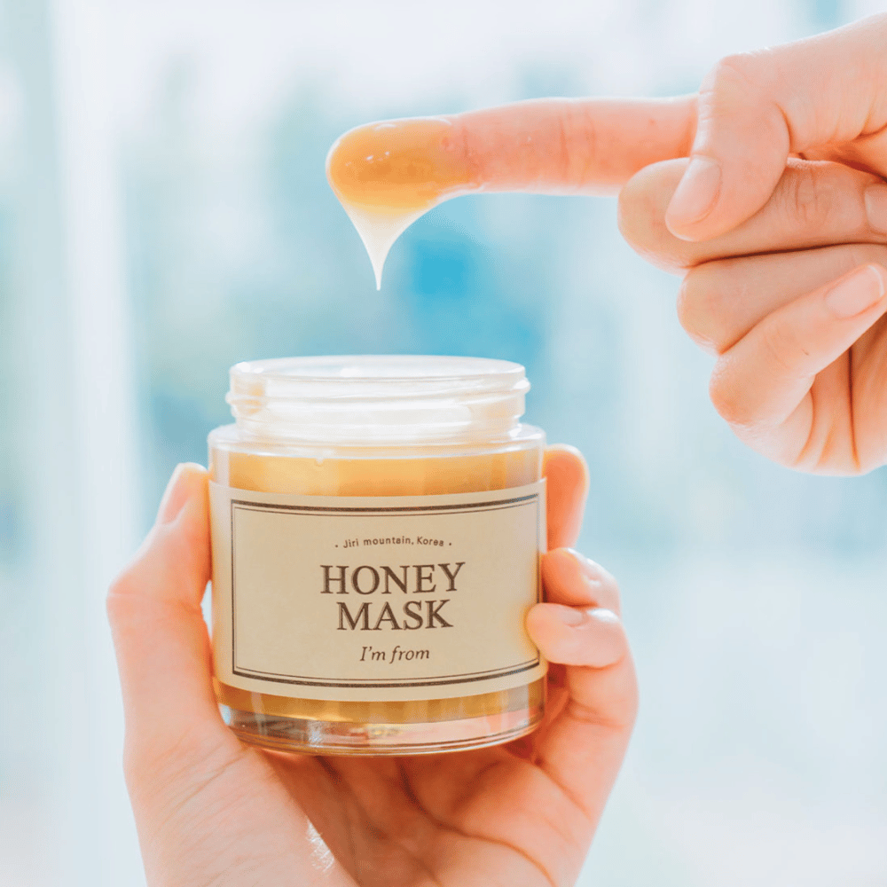 A hand holds an open jar of "I'm from Honey Mask", and a finger dips into the golden mask against a light background.