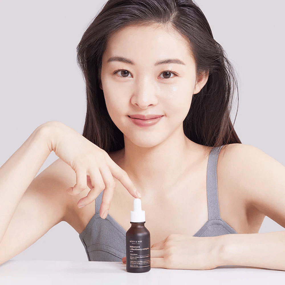 A woman with long, dark hair holds MARY & MAY Idebenone + Blackberry Complex Serum, a dark glass bottle with a dropper. She has a happy smile and points at the bottle with her other hand. The woman is dressed in a gray, sleeveless top and appears to be in a bright and simple environment, emphasizing the product's focus on skincare and beauty.
