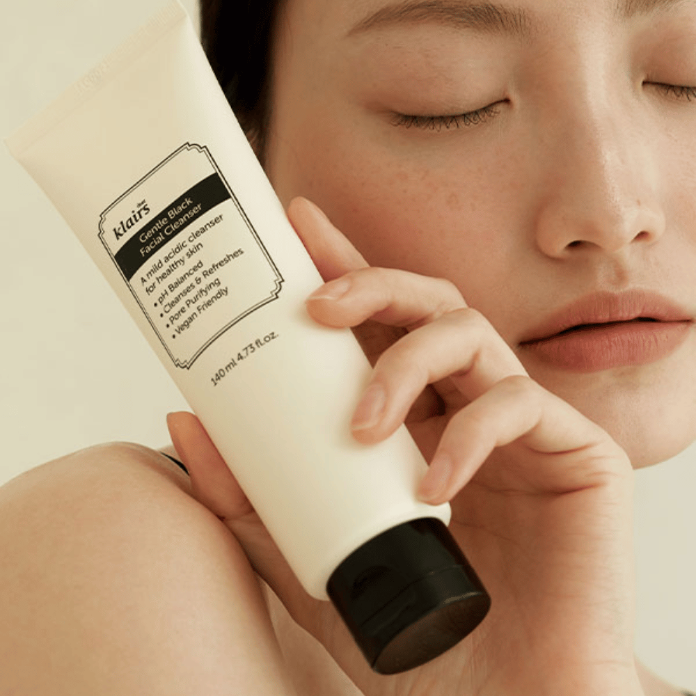 A person holds a tube of Klairs Gentle Black Facial Cleanser against their cheek, closed eyes and natural skin tone visible in the background.