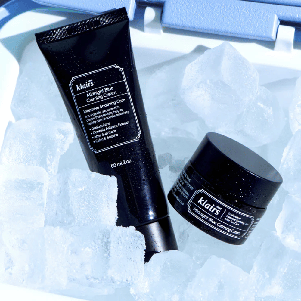 A tube and jar of Klairs Midnight Blue Calming Cream lie on ice cubes, focusing on soothing and moisturizing properties, against a blue background.