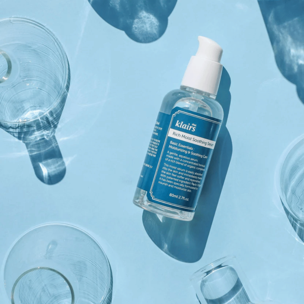 A bottle Klairs Rich Moist Soothing Serum among water glasses with reflections on a blue background.