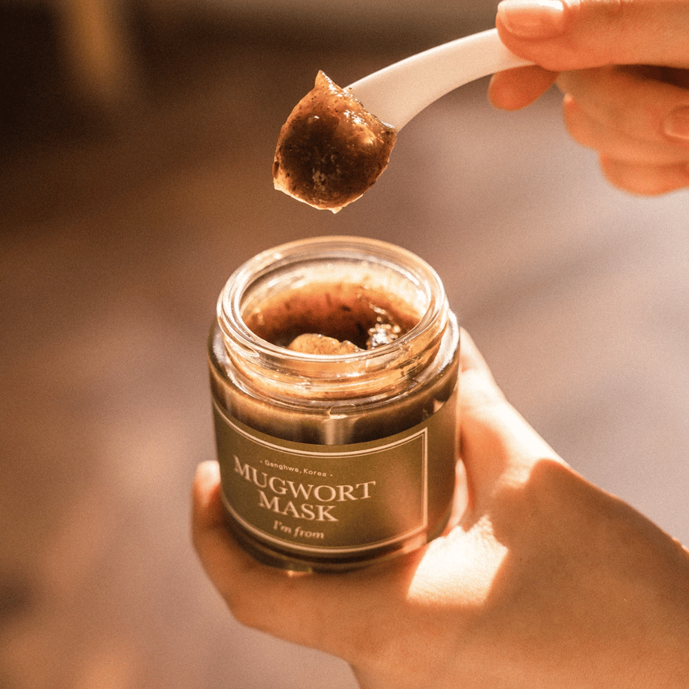 Close-up of a hand holding I'm From Mugwort Mask while a spoon lifts the herb-based gel mask out of the jar, in warm natural light.