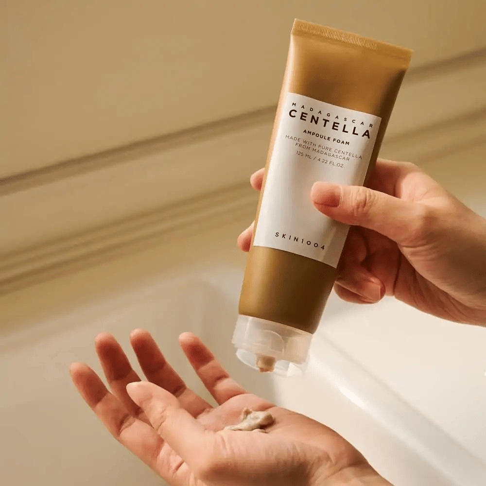 The product in the image is "Madagascar Centella Ampoule Foam" from SKIN1004. It is a facial wash in the form of a foam, made from Centella from Madagascar. It contains 125 ml / 4.22 fl.oz. A hand presses out a small amount of product for use.