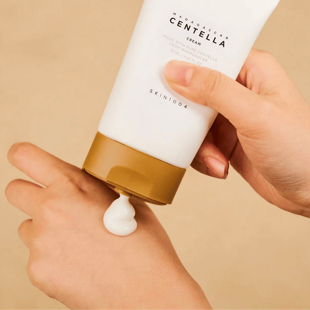 The image shows a person holding a tube of "Madagascar Centella Cream" from SKIN1004. It is a skin cream made from pure Centella from Madagascar, in the size of 75 ml / 2.53 fl.oz. The cream is applied to the hand and is intended to provide nourishment and hydration to the skin.