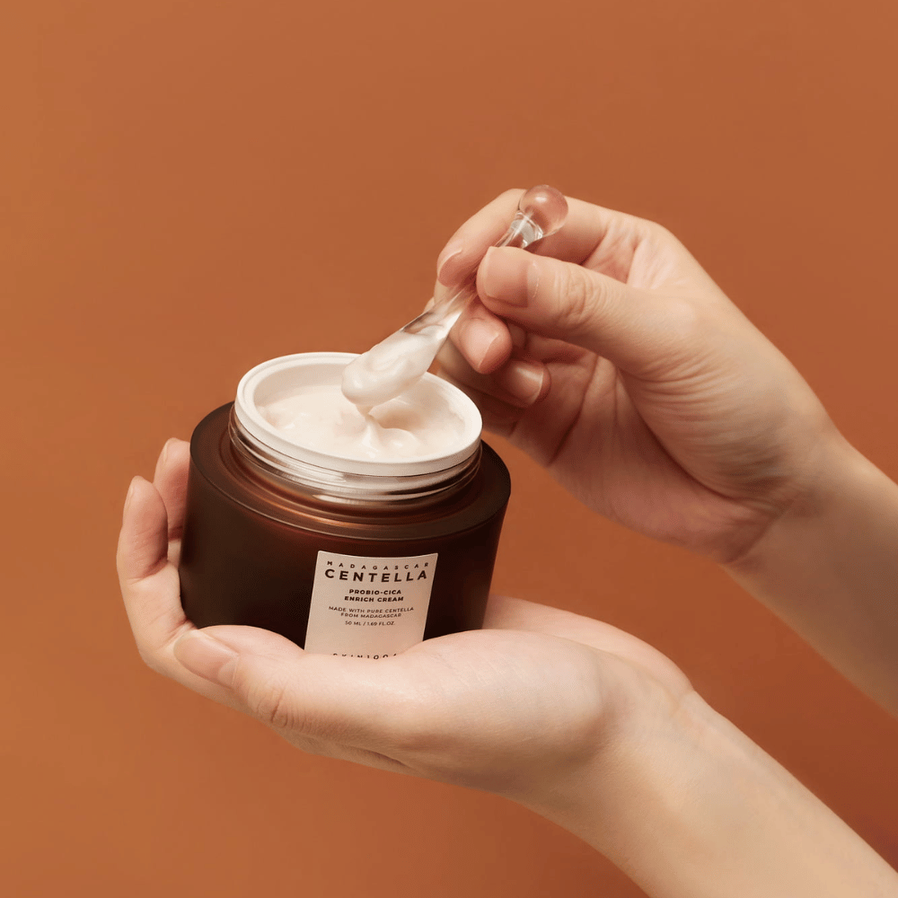 Hands holding an open jar of Madagascar Centella Probio-Cica Enrich Cream and a spatula with cream against a uniform terracotta-colored background.