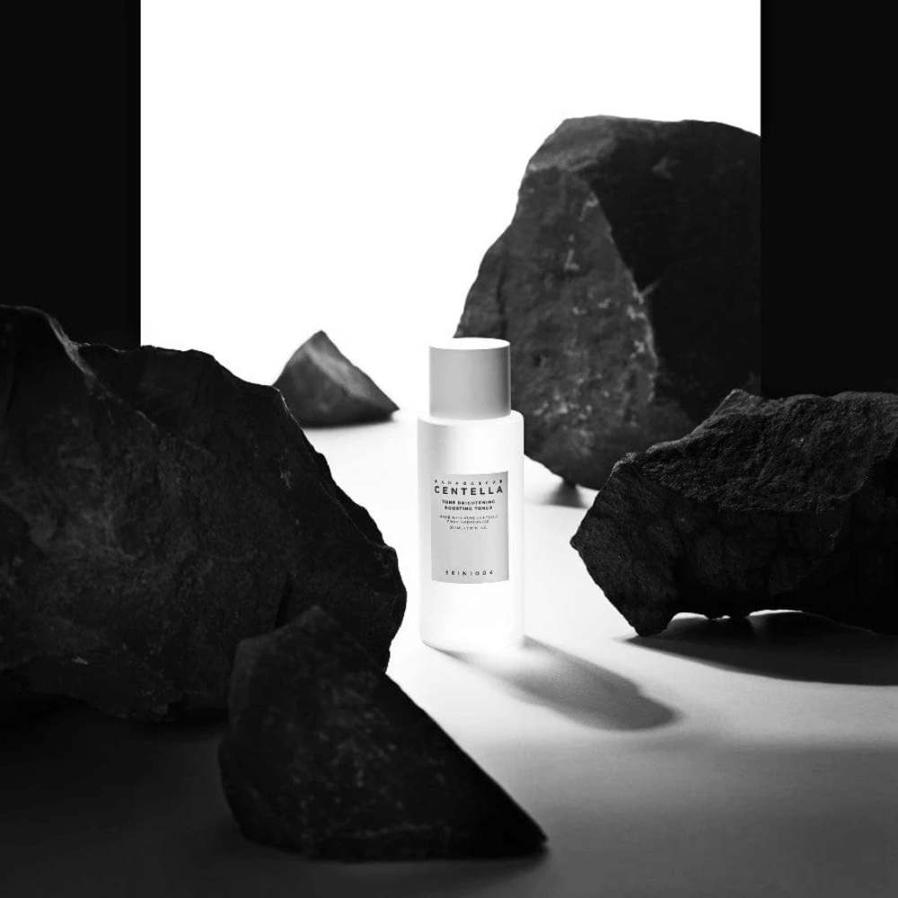 White bottle SKIN1004 Madagascar Centella Tone Brightening Boosting Toner  placed dramatically among large black stones against a white background.