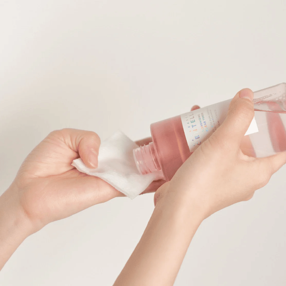 A person pours Madagascar Centella Poremizing Clear Toner from a transparent bottle onto a cotton pad. The product, which has a pink hue, is used to refresh and cleanse the skin. The hands are visible, showing the application of the toner.