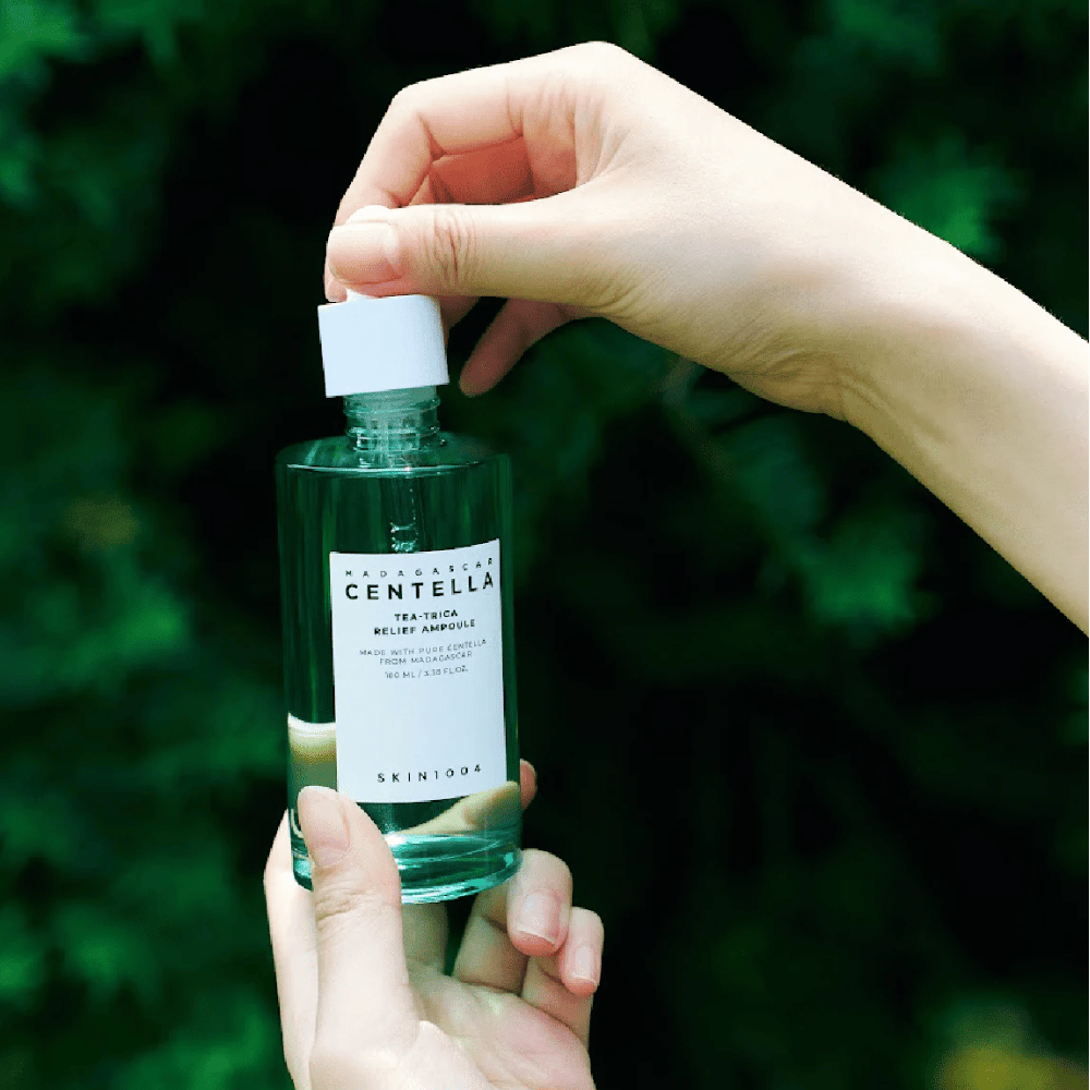 Person opens SKIN1004 Madagascar Centella Tea-Trica Relief Ampoule – green glass bottle with white label held in hands in front of lush background.