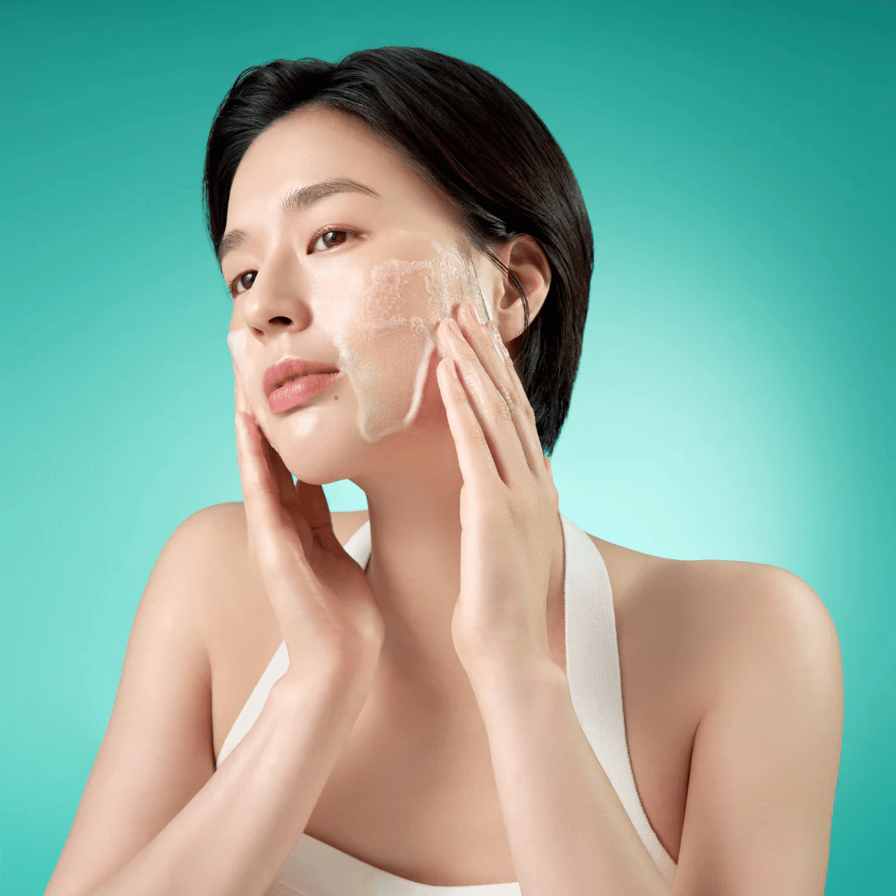 Woman cleanses her face with a mild foaming facial cleansing foam against a green background.