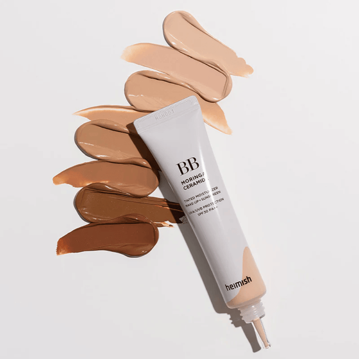 Heimish Moringa Ceramide BB Cream surrounded by several shade swatches in different skin tones. The image shows the product's wide color range and the creamy consistency that provides natural coverage and an even skin tone.
