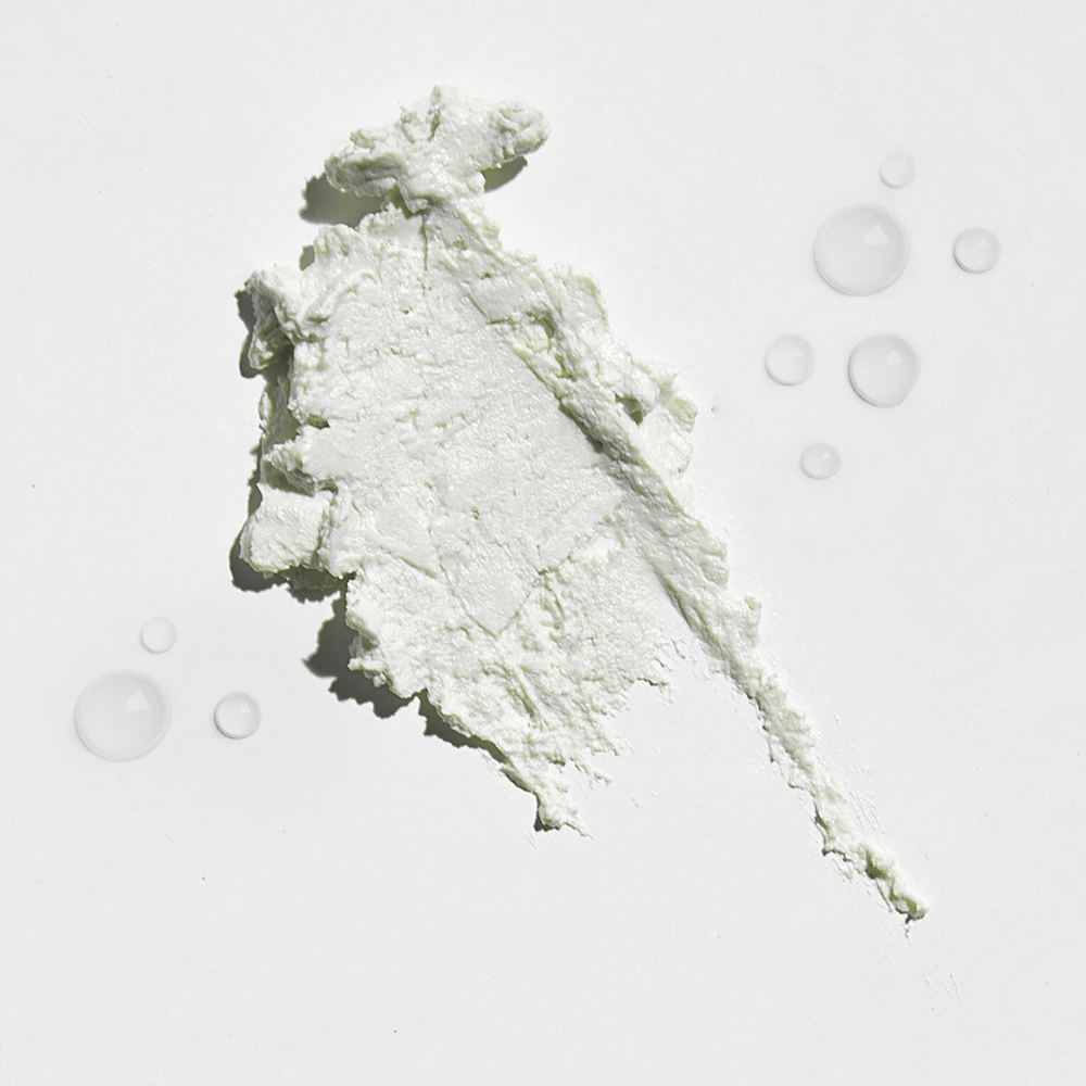 Close-up of a white, creamy substance on a white background, with some water droplets around it. The texture appears to be a sun powder or foundation with a light glow.