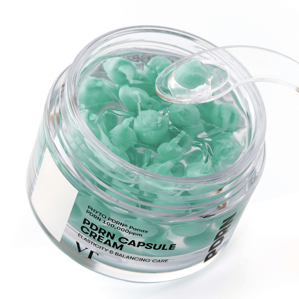 Close-up of an open jar with VT PDRN Capsule Cream. The contents consist of small green capsules in a transparent gel, with an accompanying spatula for application. The text on the jar reads "Elasticity & Balancing Care".