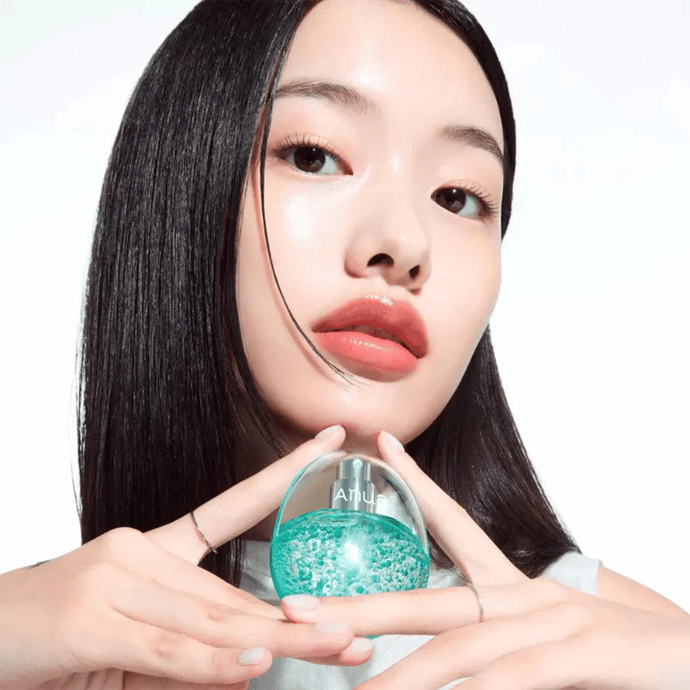 Close-up of a model holding a round, transparent Anua bottle filled with turquoise-colored capsules or gel beads. The image focuses on the skin's radiance and the product's innovative design, creating a fresh and modern impression.