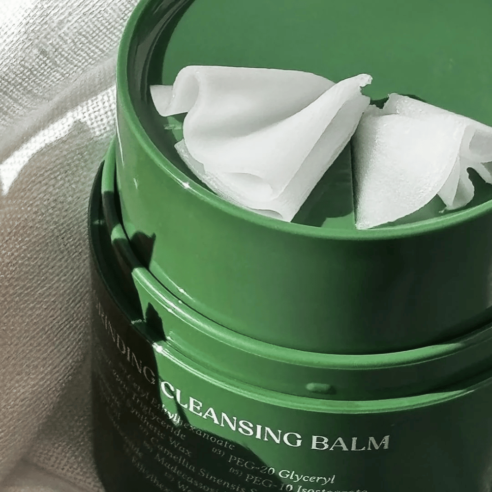 The image shows a green jar with "Cleansing Balm", which is opened to reveal the included spatula and the soft contents. The creamy texture reflects light, giving a sense of luxury and effectiveness in cleansing.