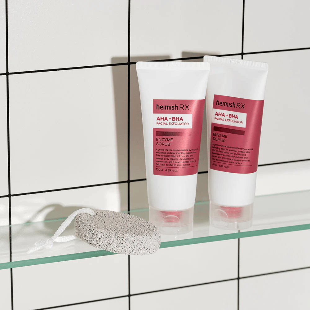 Two tubes of Heimish RX AHA + BHA Facial Exfoliator placed on a glass shelf in a bathroom with a white tiled wall. Next to the products is a pumice stone with a white string. A stylish and minimalist skincare image.