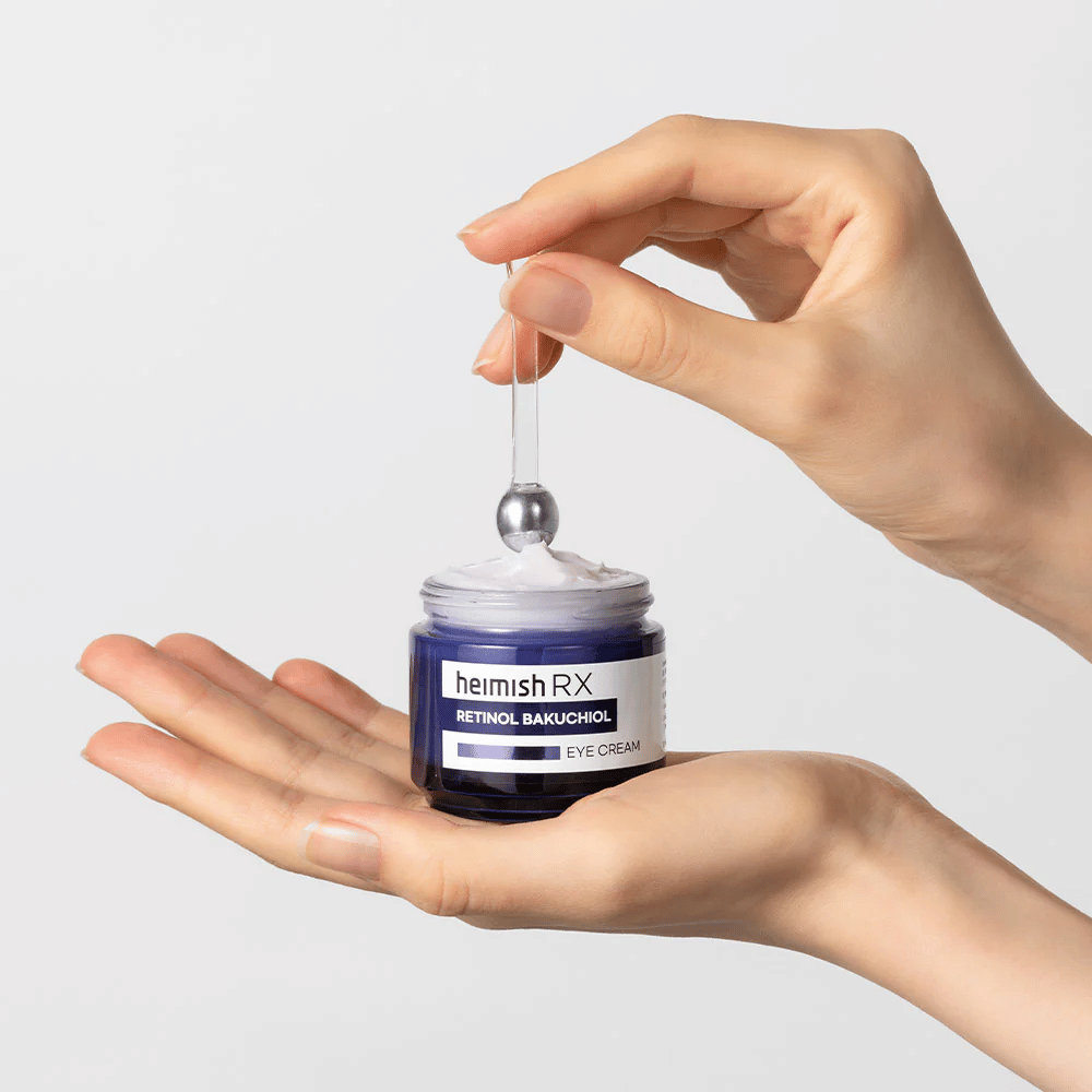 Heimish RX Retinol Bakuchiol Eye Cream in an elegant blue jar, lifted with a metal applicator. An eye cream with retinol and bakuchiol to reduce fine lines and provide firmer skin.